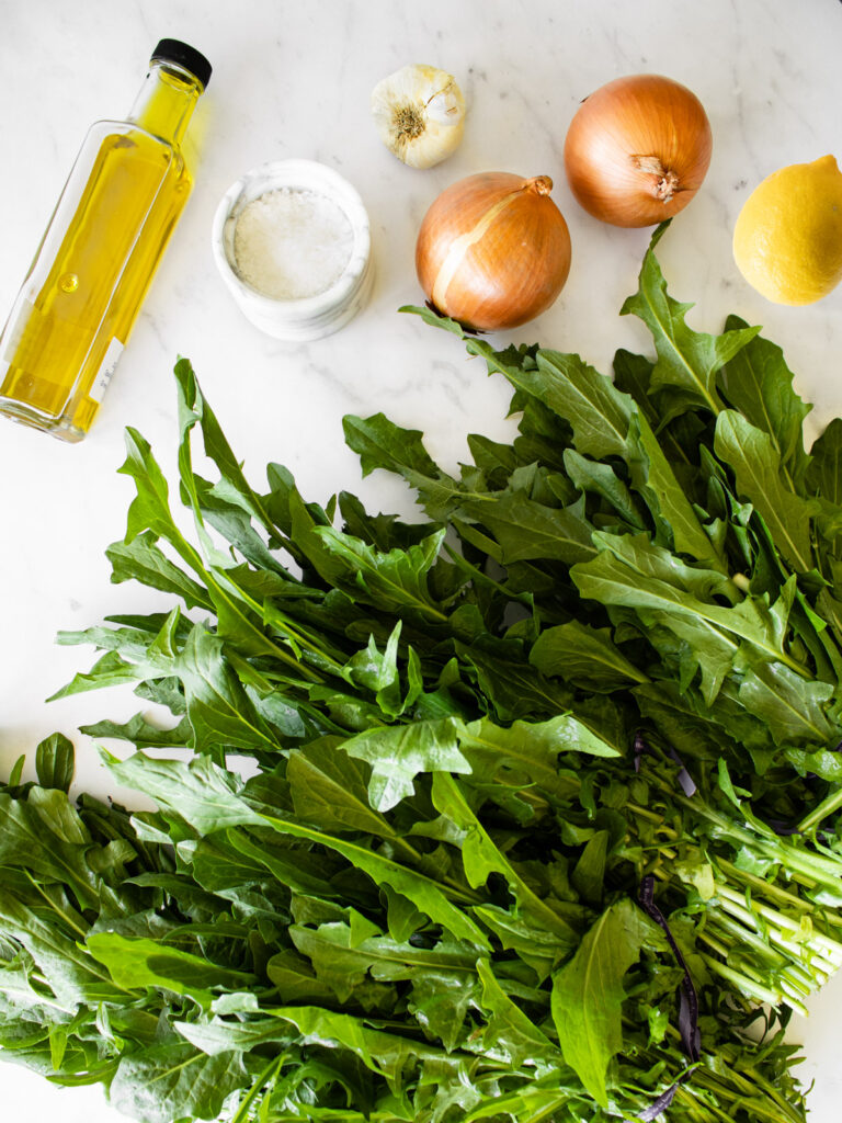 several bunches of dandelion greens, olive oil, two brown onions, one head of garlic, one lemon, and a small bowl of salt on a white marble countertop