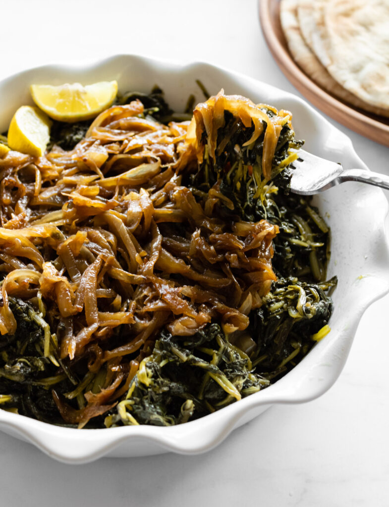 instant pot dandelion greens with caramelized onions and lemon wedges in a white ruffle ceramic dish with a fork pulling some out and pita bread in the corner