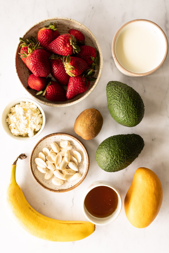 bowl of strawberries, bowl of milk, dish with peeled almonds, small bowl of honey, small bowl of ricotta, two avocados, a kiwi, a mango, and a banana on a white marble countertop