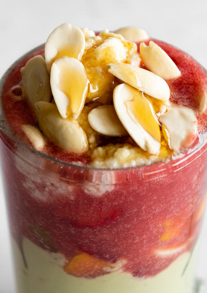 close up of the glass showing avocado smoothie layer, fruits, strawberry smoothie layer, cream, almonds, and honey