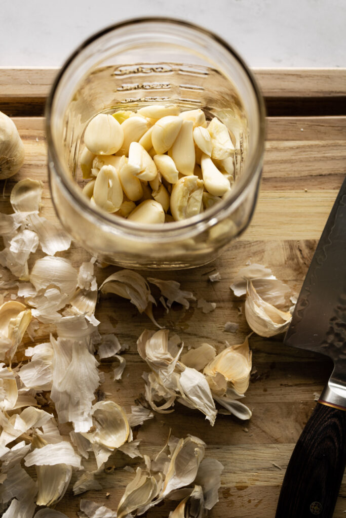 Cutting board with garlic peel, knife, and peeled garlic in a jar