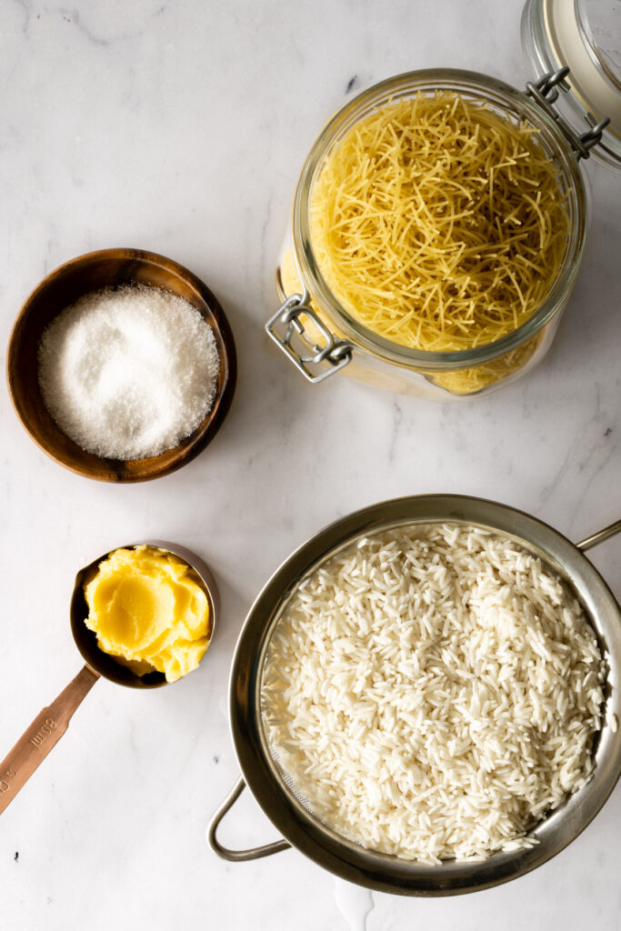overhead ingredient shot including rice in a strainer, ghee in a measuring cup, vermicelli in a jar, small bowl with salt, all on a white marble countertop