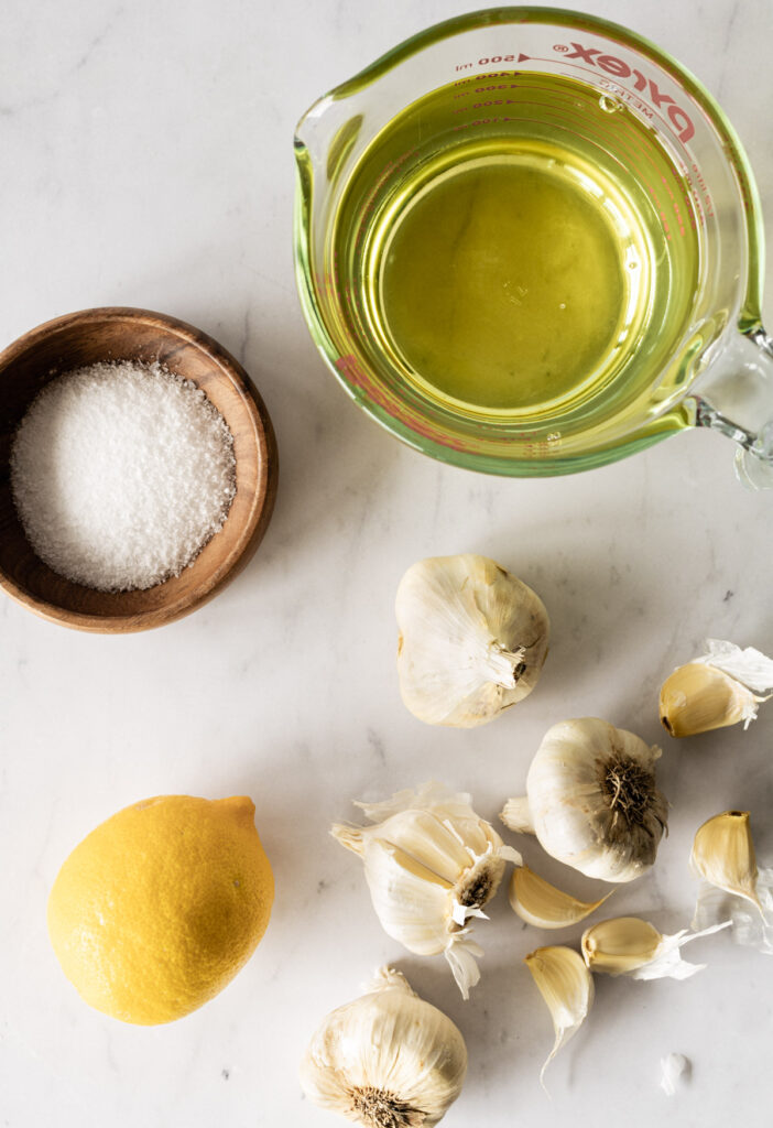 A few garlic heads, a whole lemon, a small dish with salt, and a measuring cup with oil