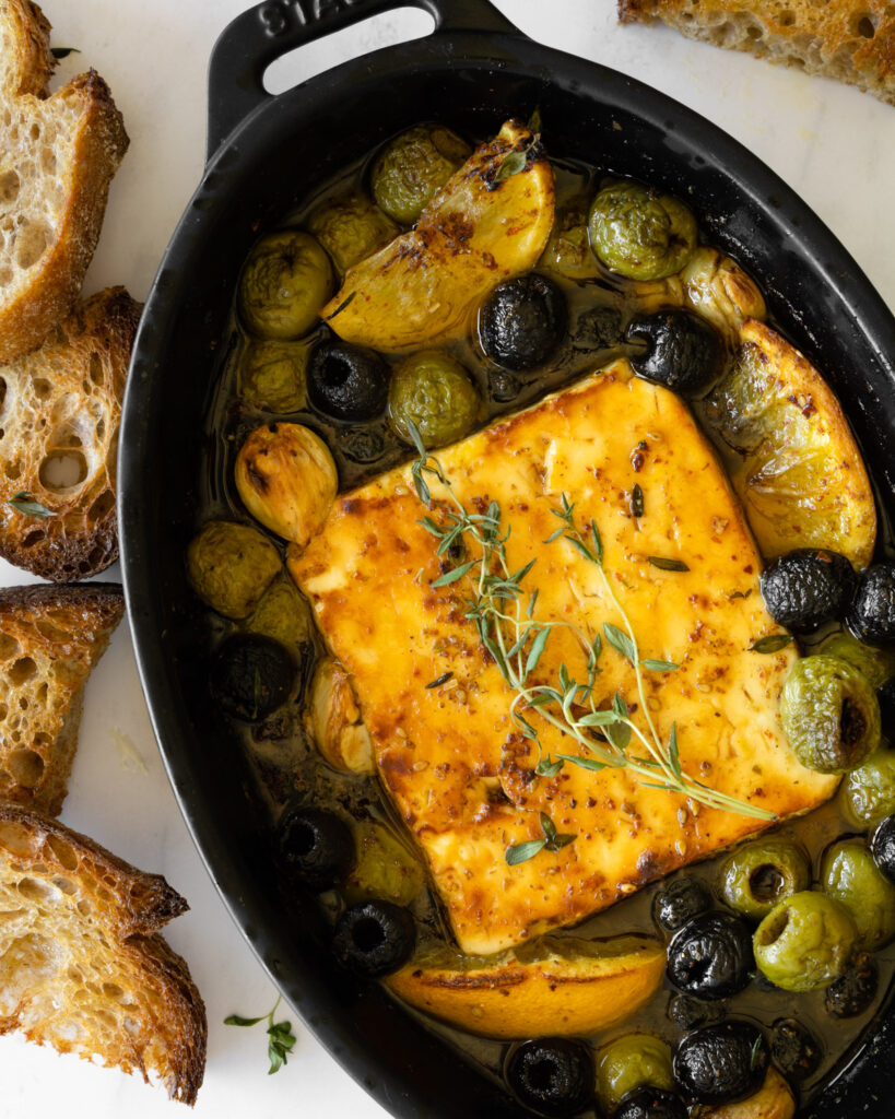 baked feta with olives and honey garnished with fresh thyme and bread on the side.
