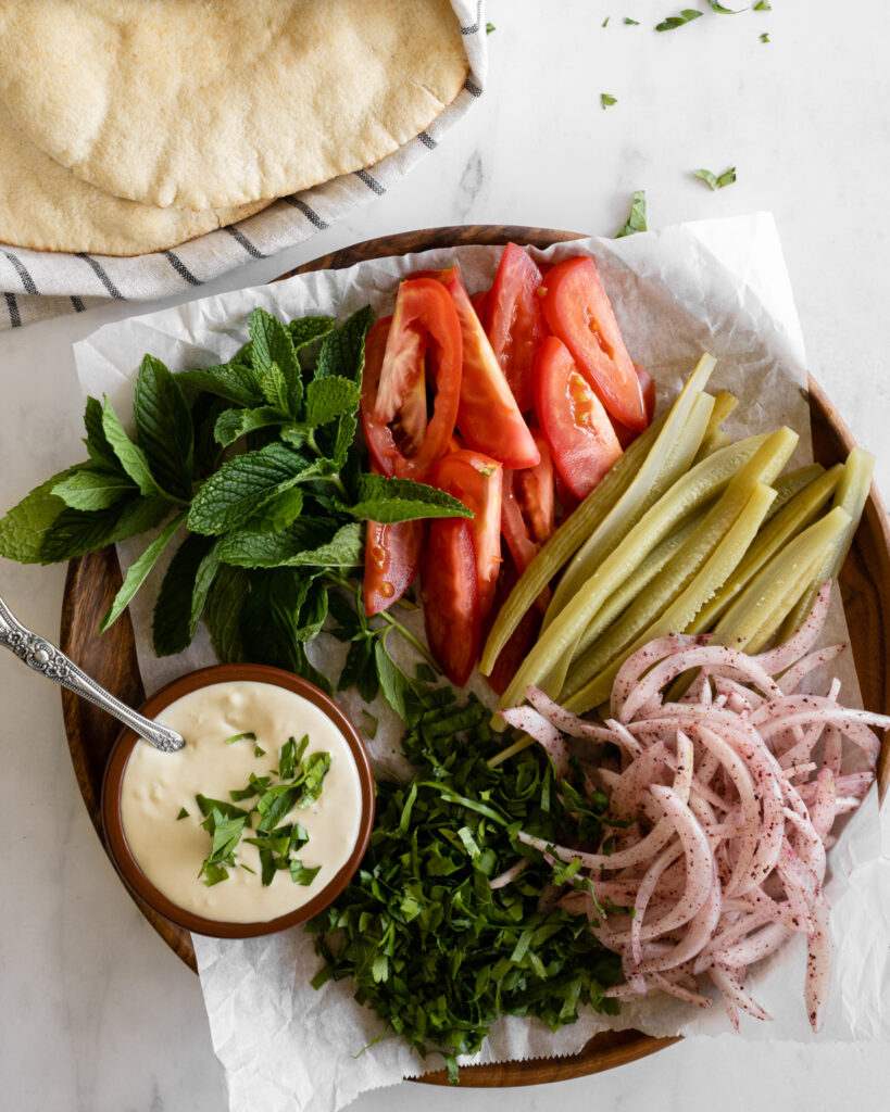 shawarma toppings and tahini sauce arranged on a wooden platter with pita bread on the side.