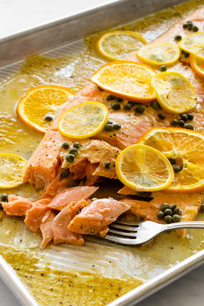 slow roasted salmon with citrus and capers on a baking sheet with a fork flaking the salmon.