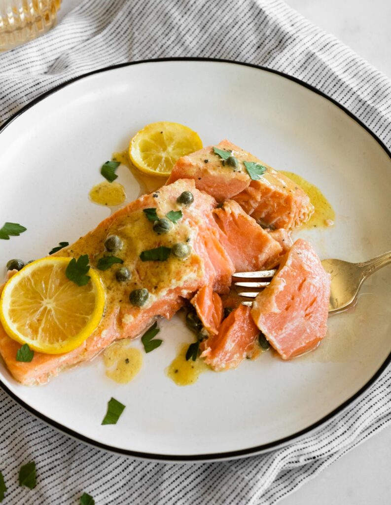 slow roasted salmon with citrus and capers on a white plate with a fork flaking the salmon.