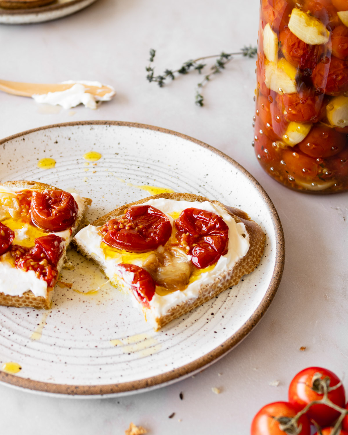 cherry tomato confit and garlic confit on toast with labneh on a ceramic plate with a jar in the background.