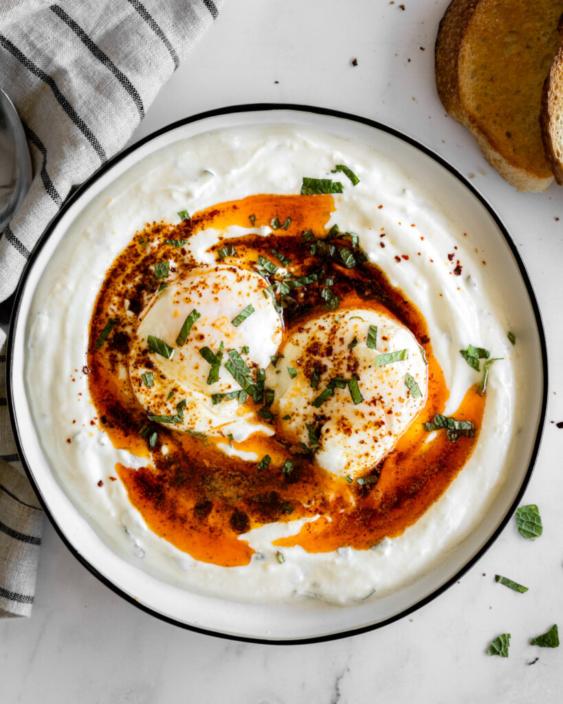 turkish eggs (cilbir) on a white plate with bread on the side.