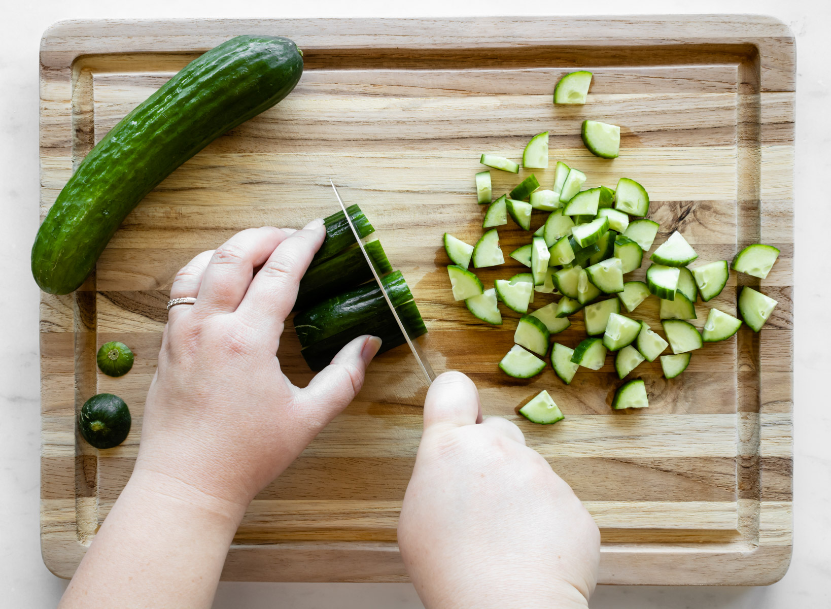hands chopping cucumbers with a knife on a wooden cutting board.