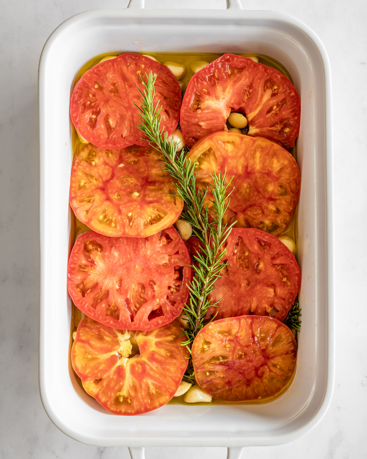 sliced tomatoes, garlic, and rosemary arranged in a white baking dish.