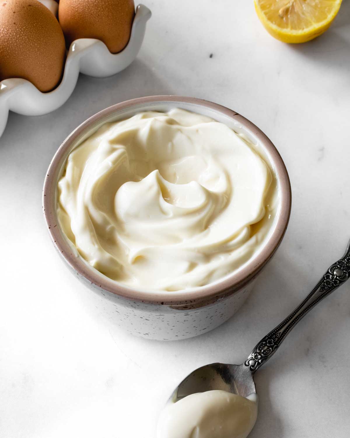 homemade mayonnaise in a small ceramic bowl with eggs and a lemon and a spoon on the counter.