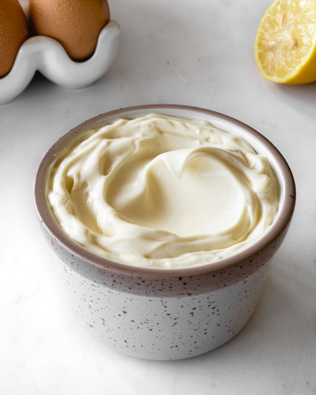 homemade mayonnaise in a small ceramic bowl with eggs and a lemon in the background.