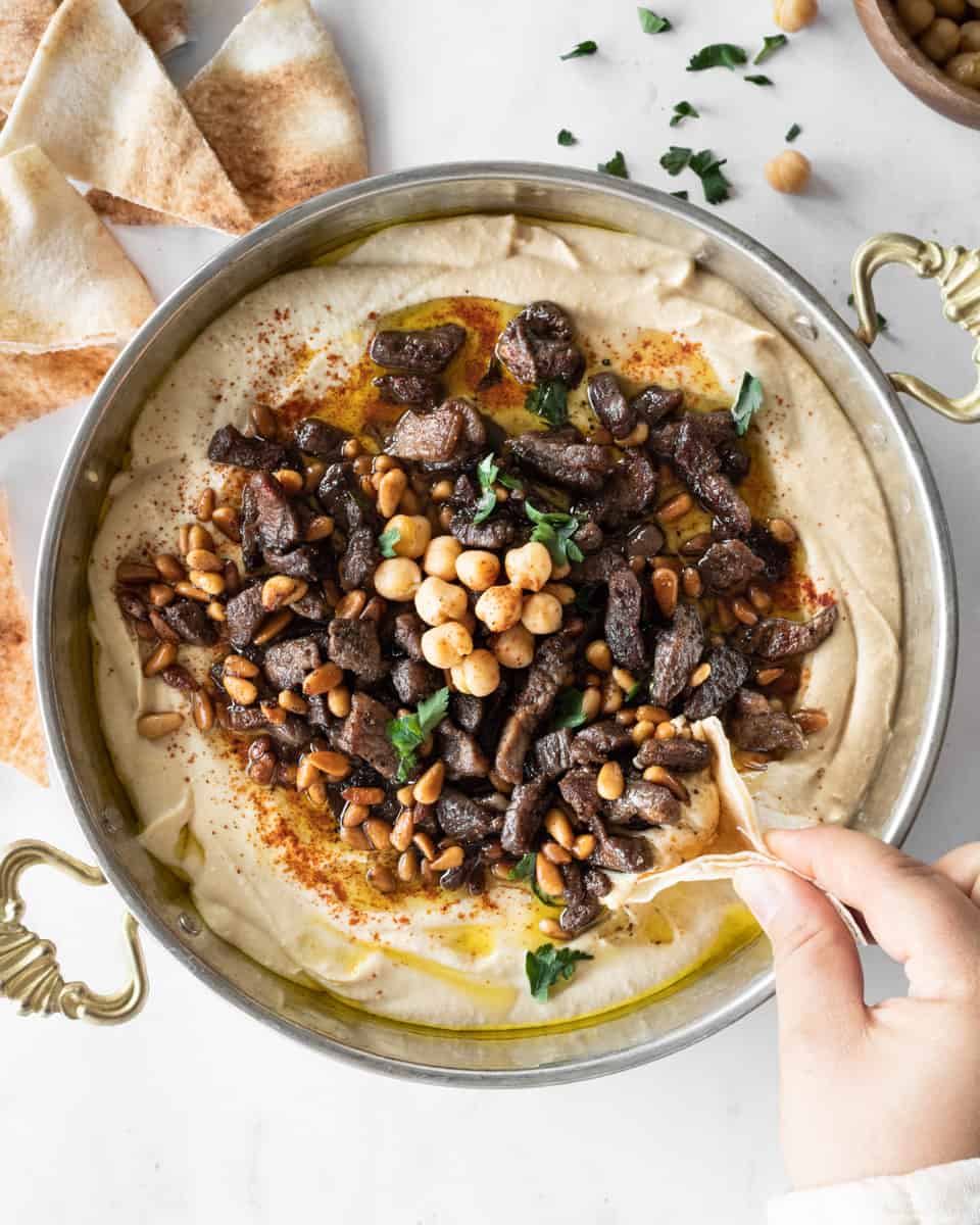 a hand holding pita bread and scooping up hummus with meat.