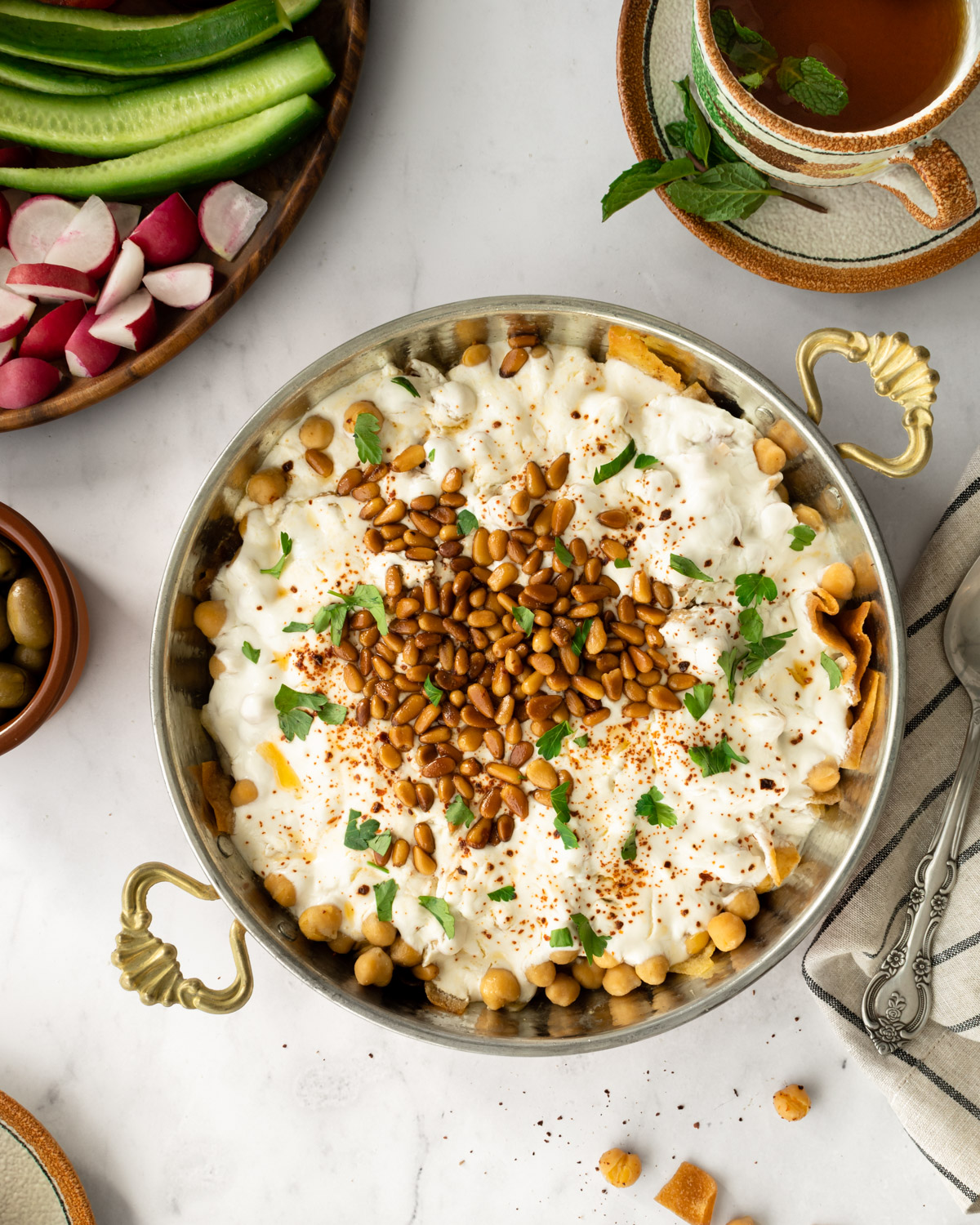 chickpea fatteh in a bowl surrounded by fresh veggies, tea, and a cloth napkin.