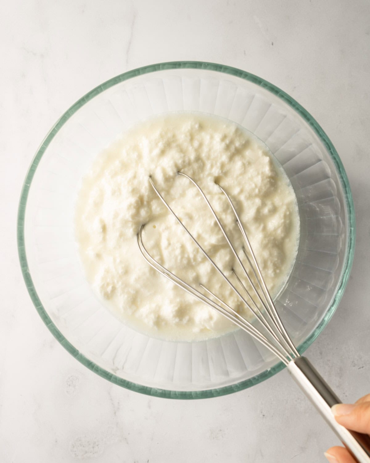 yogurt in a glass bowl with a hand holding a whisk in it.