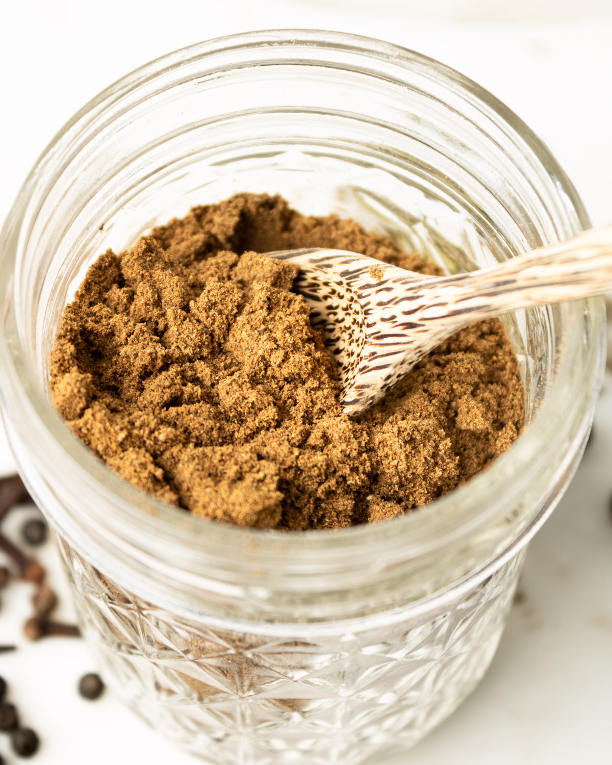 close up of Lebanese seven spice in a glass jar with a wooden spoon in it.