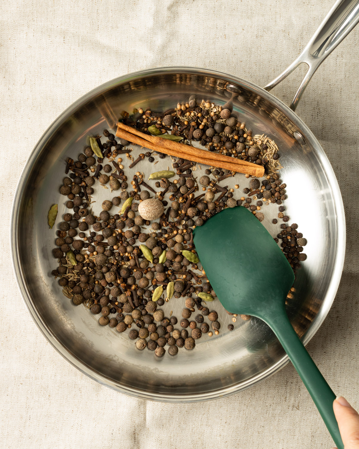 whole spices in a stainless steel pan with a spatula stirring them.