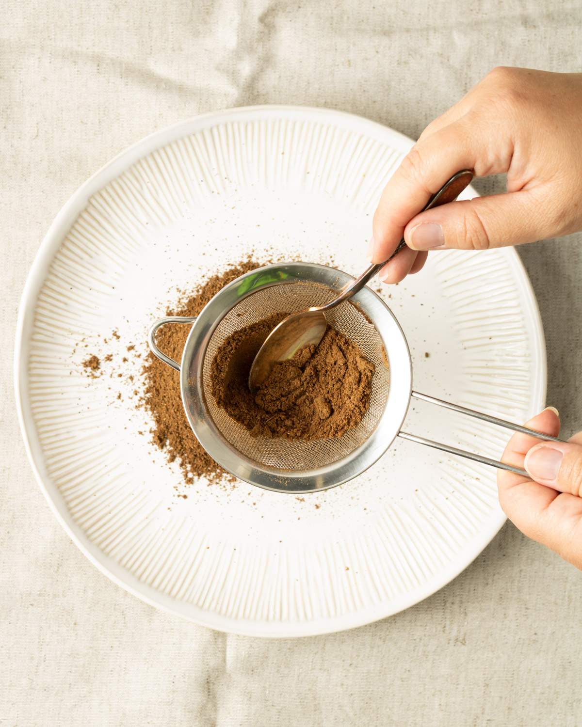 hands holding a fine mesh sieve to sift the ground spices on a plate.